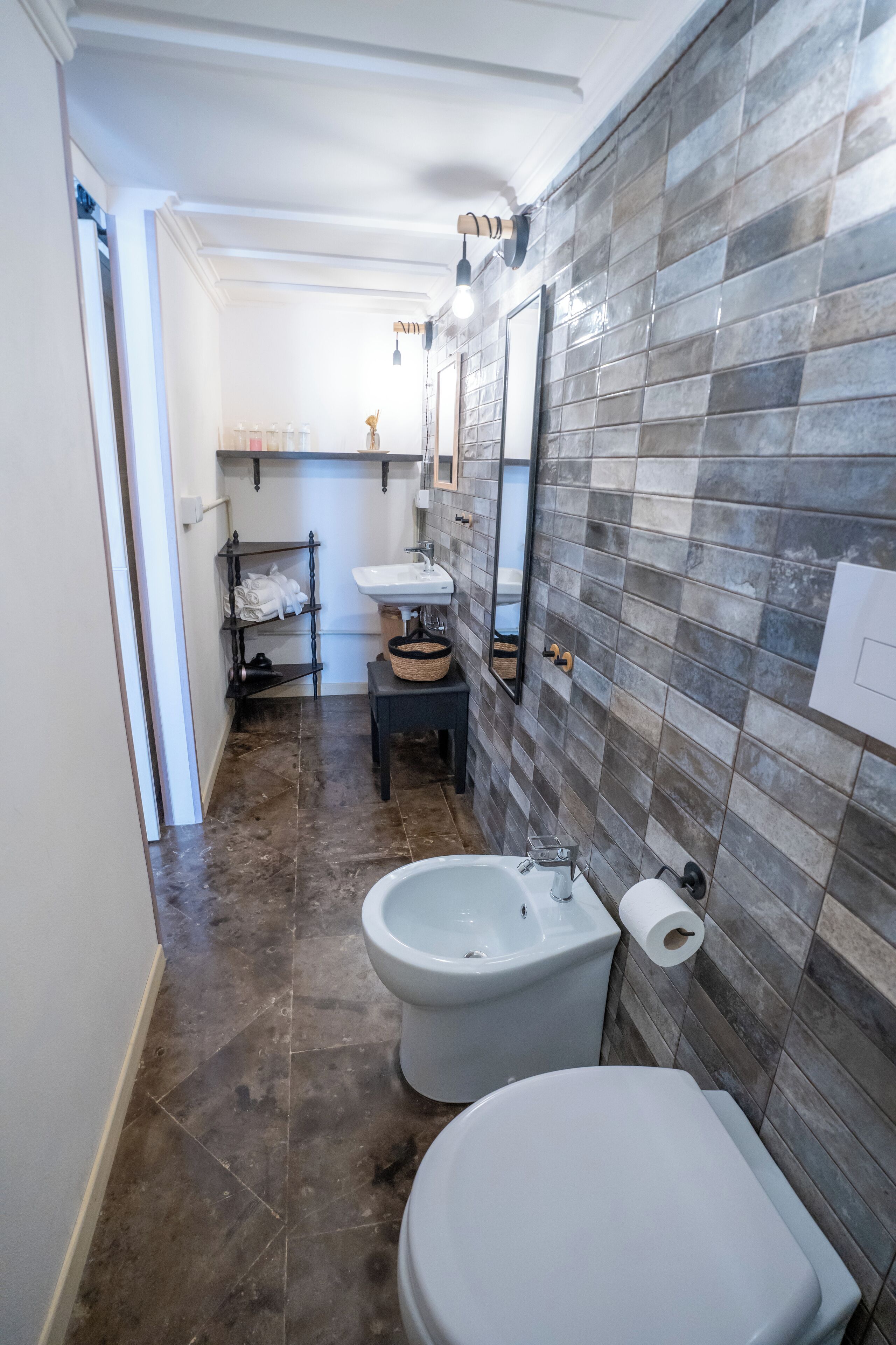Un baño con inodoro, bidé y lavabo. Hay una repisa con toallas y una cesta. Las paredes están cubiertas de azulejos con diseño de piedra.