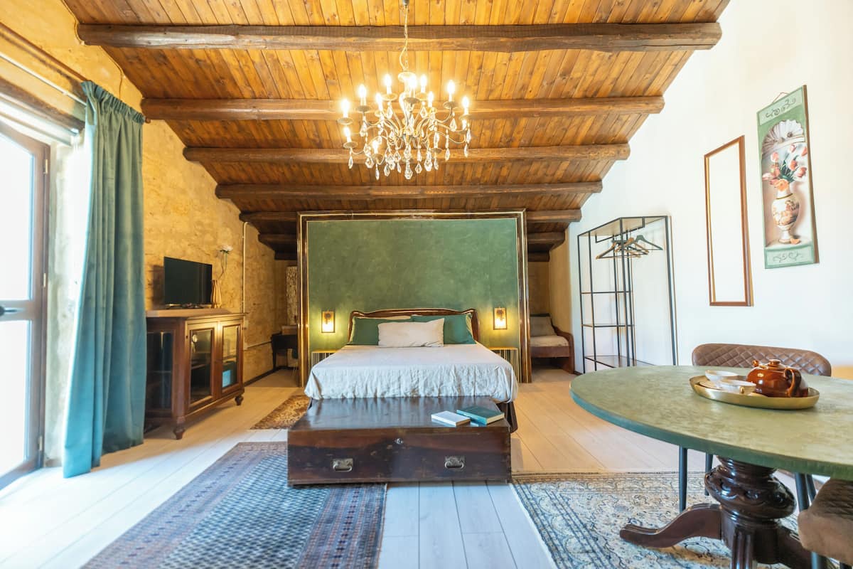 A bedroom with a wooden ceiling, a bed with green headboard, bedside lamps, a dresser, a television, and a table with a teapot.