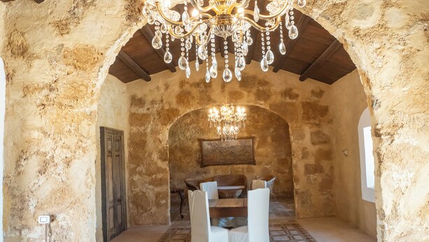 Una habitación rústica con paredes de piedra, techo de madera y un candelabro. Hay una mesa con sillas y una alfombra estampada.