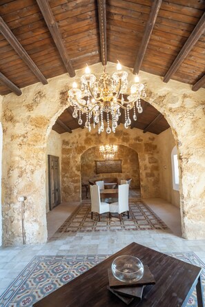 A rustic stone-walled room with a wooden ceiling and a chandelier. There is a table with chairs and a patterned rug. - Feudo Nobile (Gela)