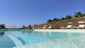 Una piscina azul cristalina con sillas de descanso blancas y un muro de piedra al fondo.