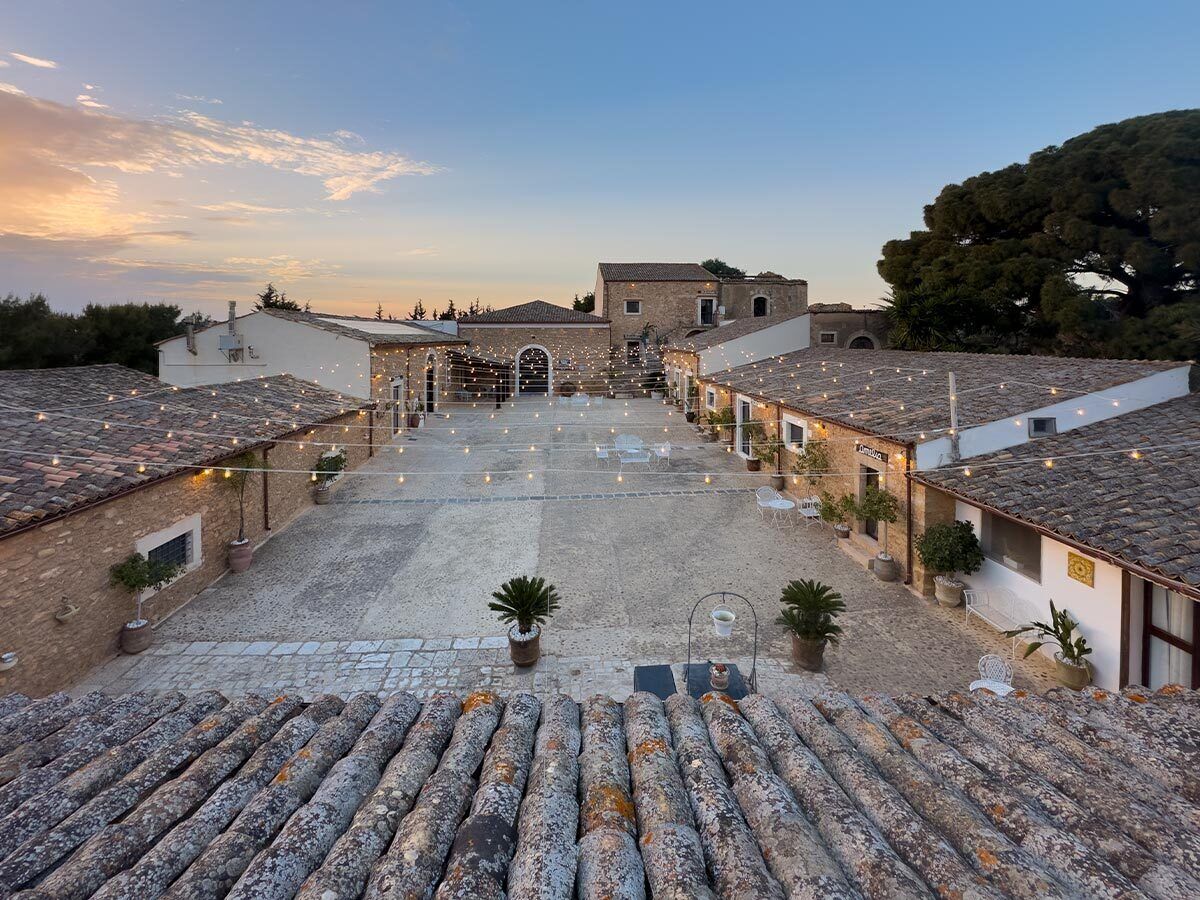 A courtyard with stone flooring, potted plants, and buildings with tiled roofs.