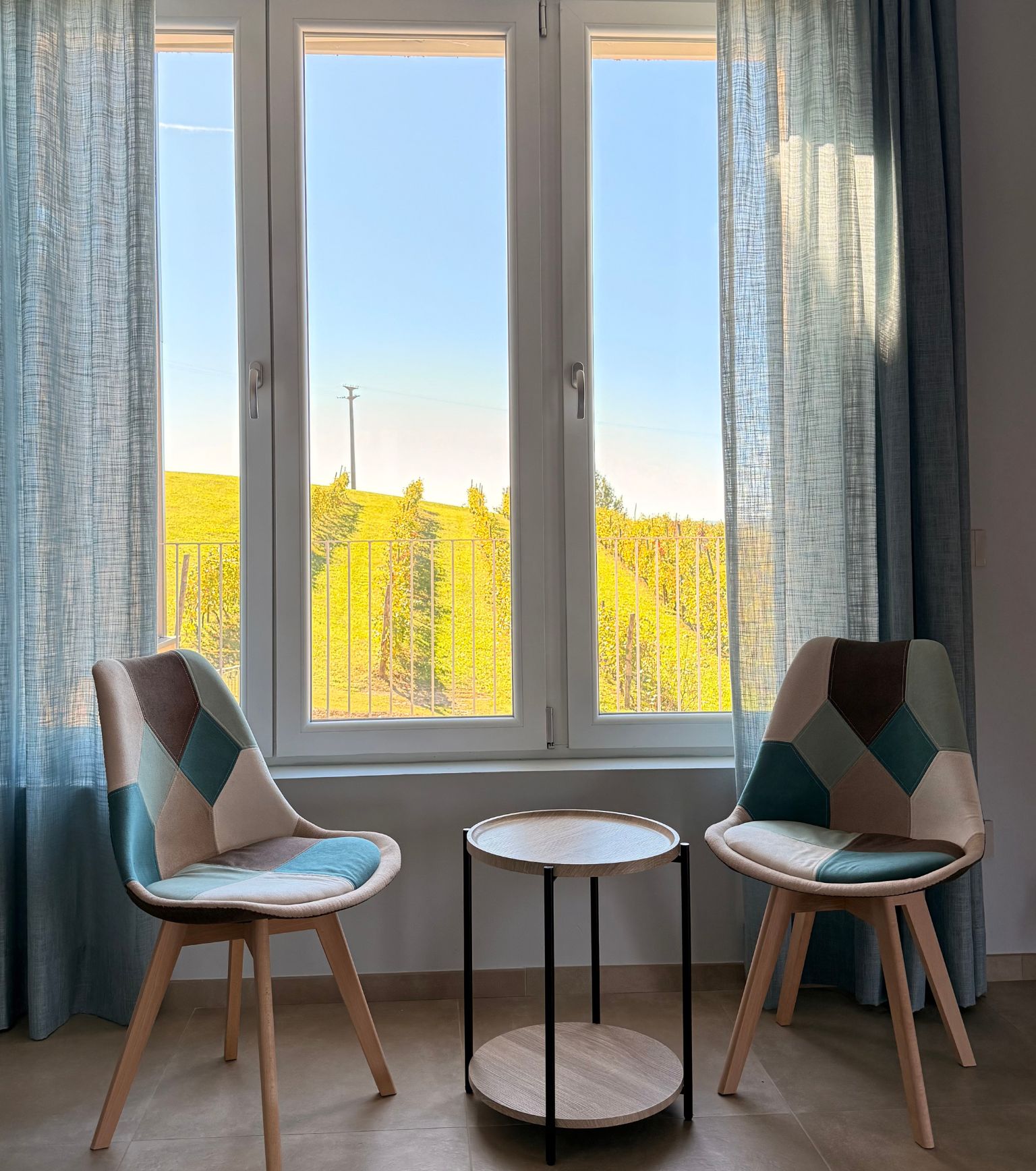 A room with two chairs and a small table, large windows with curtains, and a view of a yellow field.