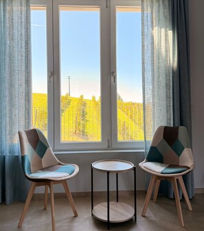 Una habitación con dos sillas y una pequeña mesa, ventanas amplias con cortinas, y vistas a un campo amarillo.