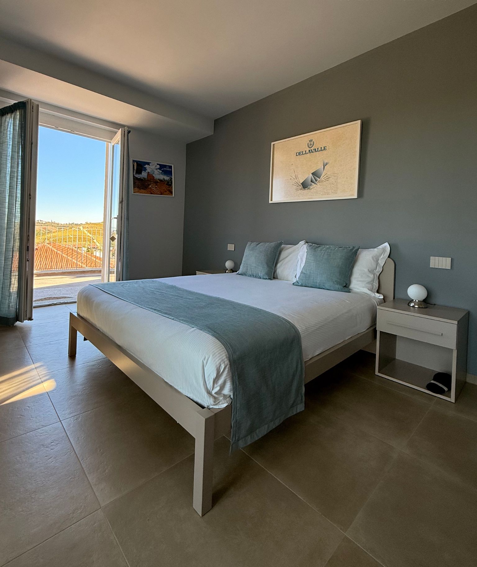 Un dormitorio con cama, mesitas de noche, un cuadro enmarco y vistas a través de un ventanal.