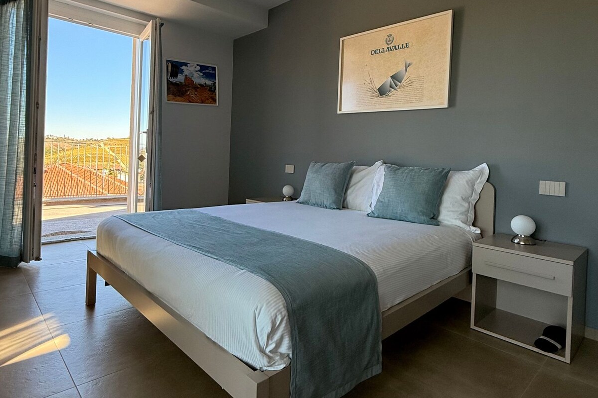 Un dormitorio con cama, mesitas de noche, un cuadro enmarco y vistas a través de un ventanal.