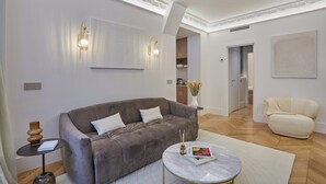 A modern living room with a grey sofa, a round coffee table, and a beige armchair. There is a bookshelf with books and decorative items, and a vase with flowers on the side.