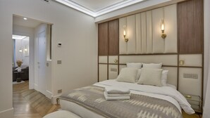 A modern bedroom with a large bed, a headboard with built-in lighting, and a visible door leading to another room.