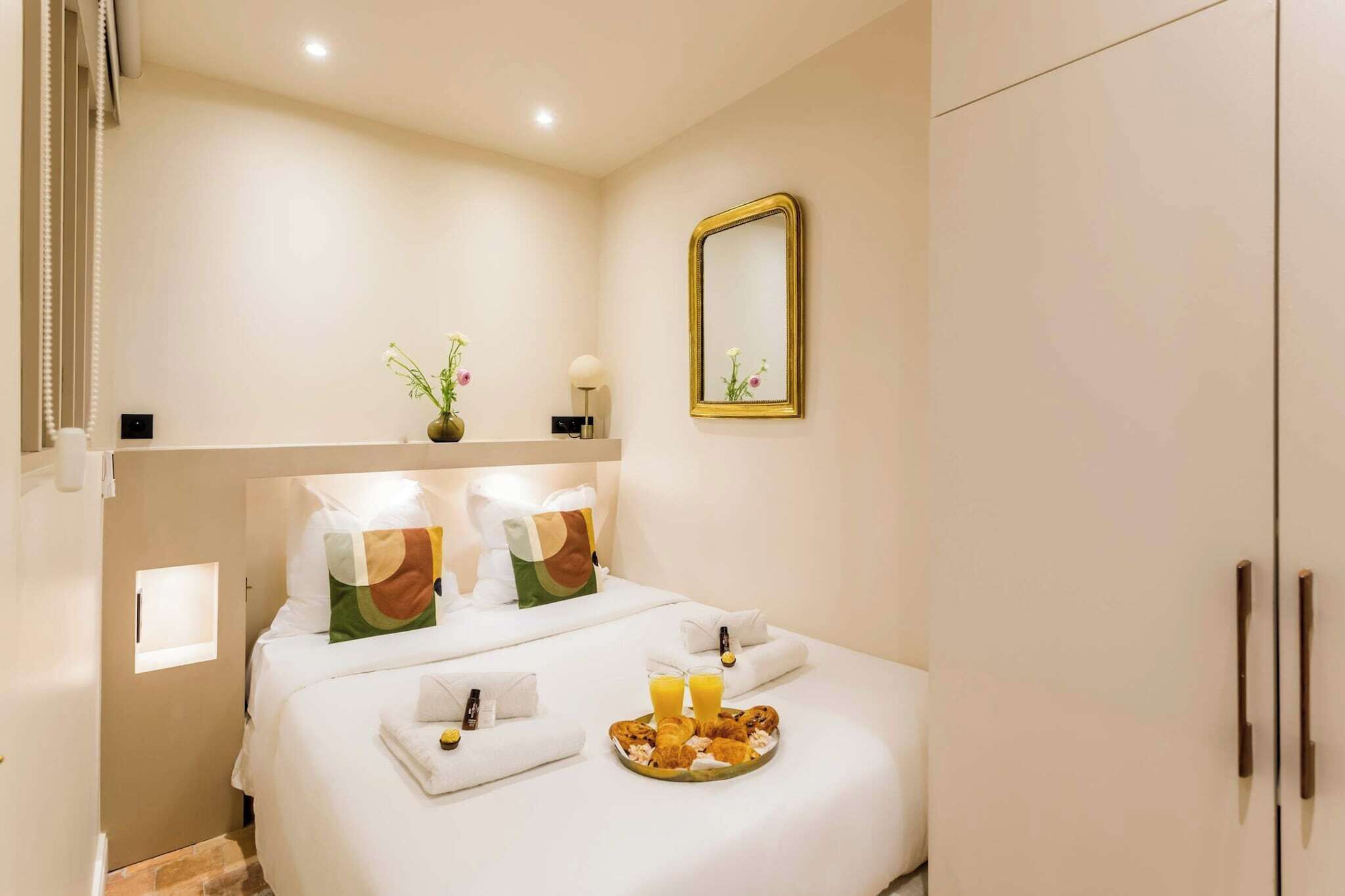 A hotel room with a neatly made bed, a bedside table with a lamp, a tray of breakfast items, and a large mirror on the wall.