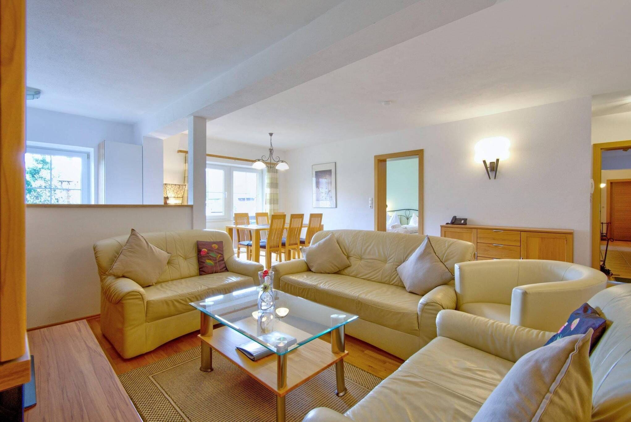A living room with a glass coffee table, two sofas, a dining area with wooden chairs, and a wooden cabinet.