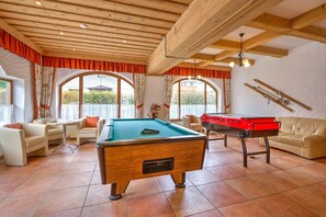 A cozy indoor space with a pool table, a foosball table, and a billiards table. There are comfortable seating areas and large windows offering views outside.