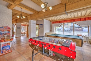 A room with a foosball table, a pool table, and a billiards table. There are wooden beams on the ceiling and a view of the outside through a large window.