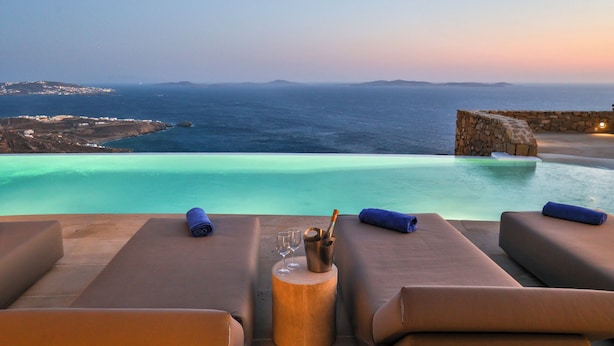 Ein Infinity-Pool mit Blick auf das Meer und entfernte Inseln, Liegestühle und ein kleiner Tisch mit Flasche und Gläsern.