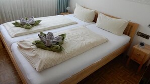 Een tweepersoonsbed met een witte spreideken en kussens, een houten hoofdbord, een nachtkastje met een lamp en een raam met gordijnen.