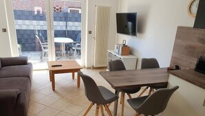 Een woonkamer met een bank, een houten tafel en vier stoelen. Er is een televisie aan de muur en een raam met uitzicht op het omliggende landschap.