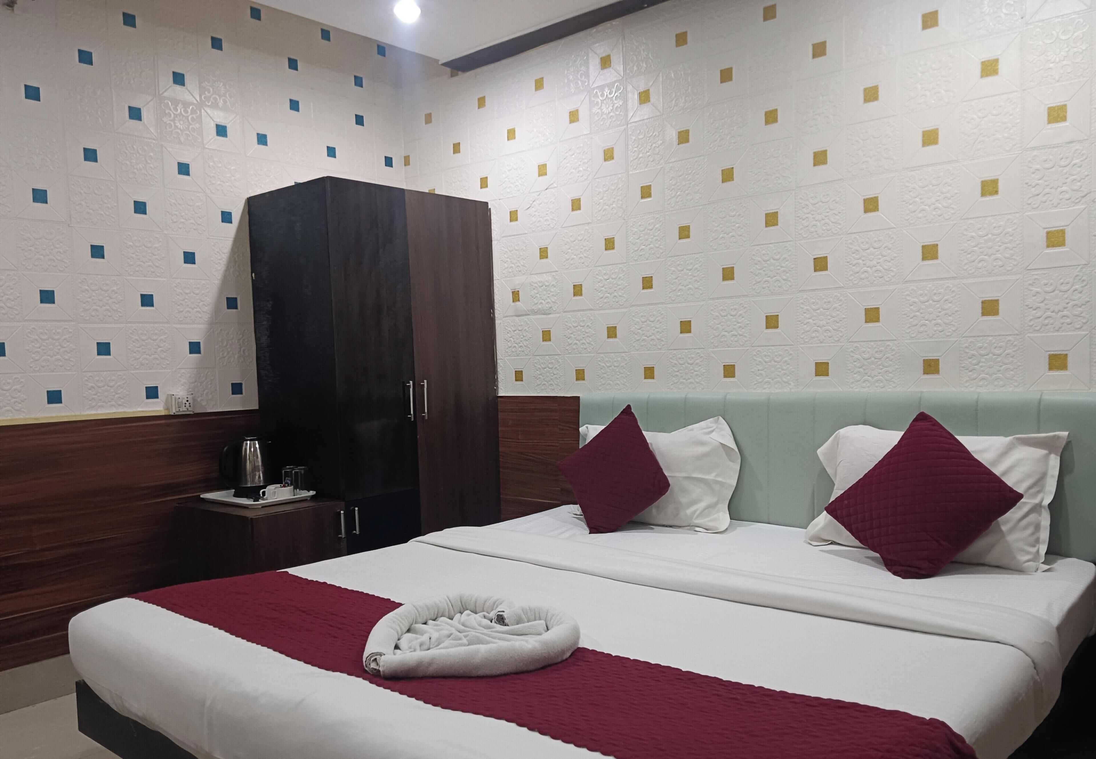 Quarto de hotel com uma cama, duas almofadas, cabeceira, guarda-roupa, mesa de cabeceira com chaleira e parede com padrão geométrico.