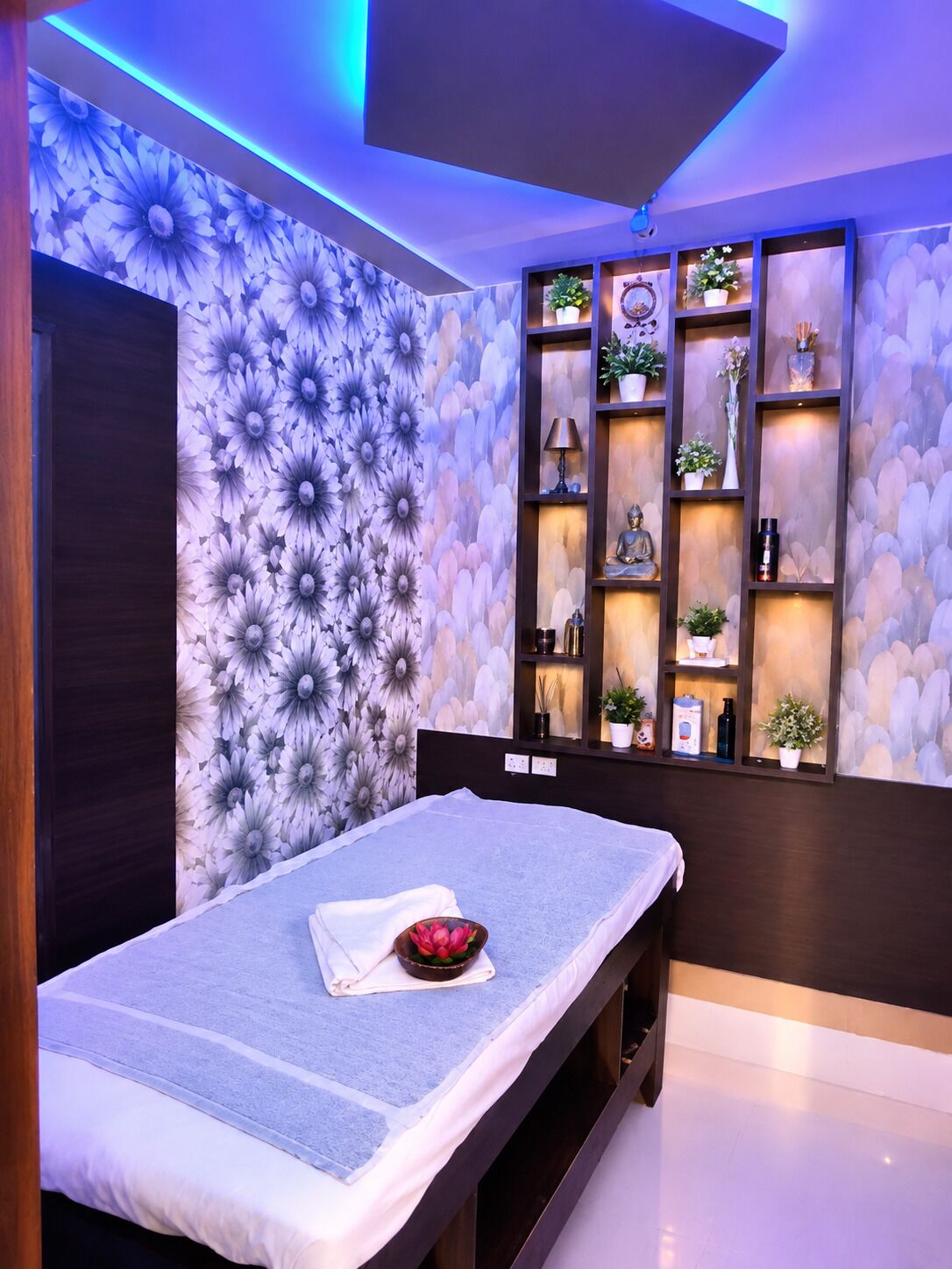 Una sala de masajes con una camilla, una pared con diseño floral y estantes con diversos objetos.