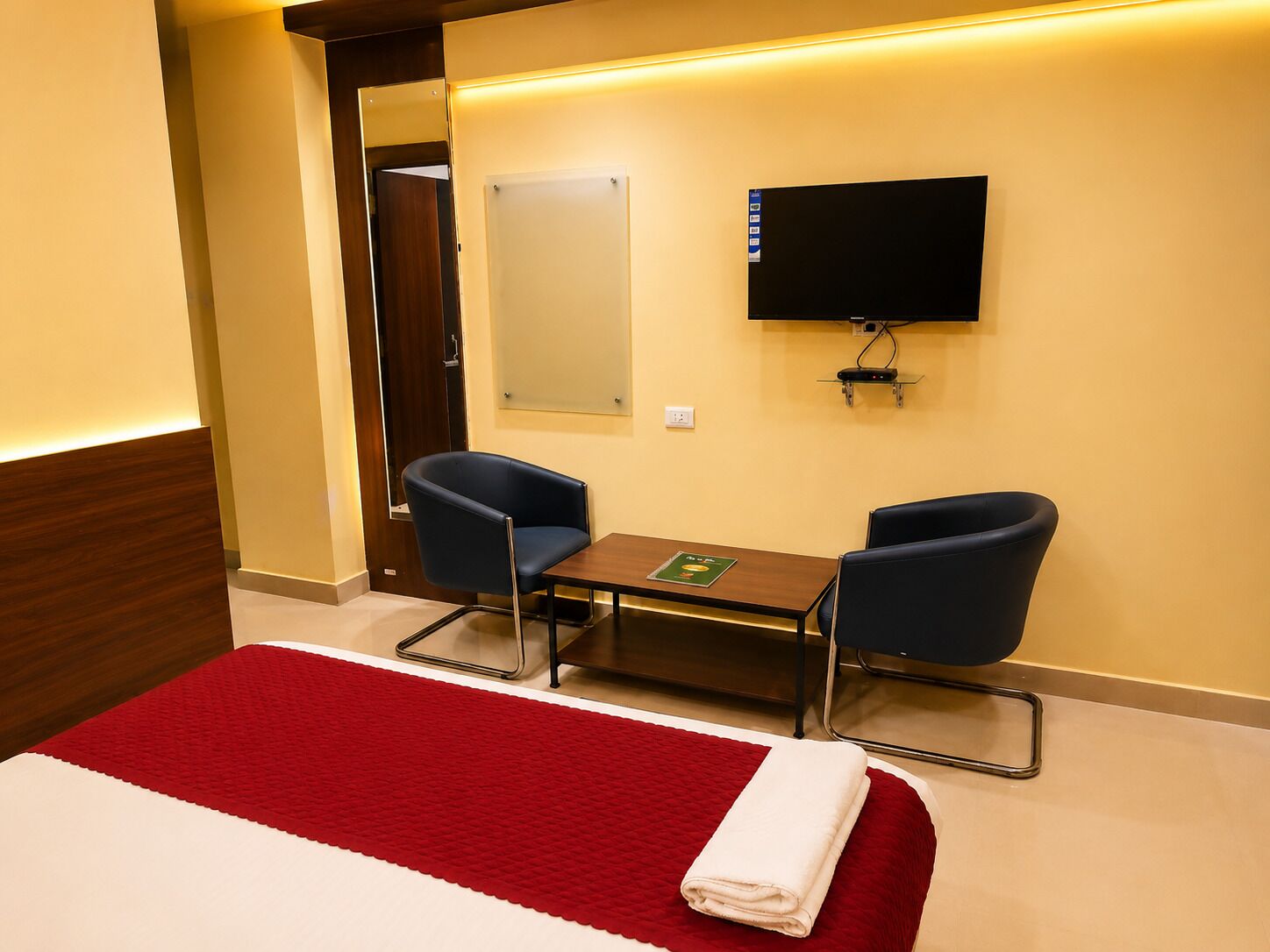 A hotel room with a bed, two chairs, a small table, a flat-screen TV mounted on the wall, and a red and white bedspread.