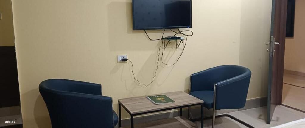 Une pièce avec deux chaises bleues, une petite table sur laquelle se trouve un livre, un téléviseur à écran plat fixé au mur et une porte sur le côté droit.