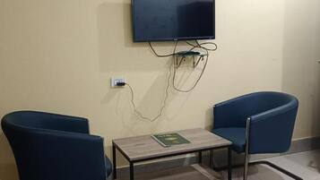 Une pièce avec deux chaises bleues, une petite table sur laquelle se trouve un livre, un téléviseur à écran plat fixé au mur et une porte sur le côté droit.