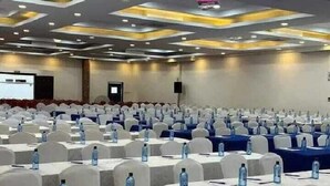 Ein Konferenzraum mit Reihen Tische, an jedem Tisch eine Wasserflasche und ein Notizblock. Der Raum zeichnet sich durch ein modernes Decken-Design mit eingelassener Beleuchtung aus.