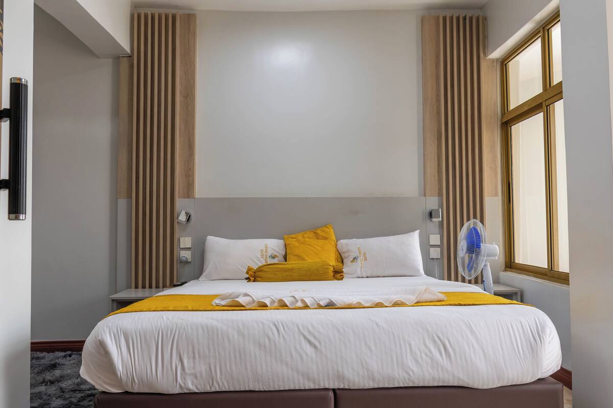 Una cama bien hecha con ropa blanca y un plaid amarillo, flanqueada por paneles de madera verticales, una mesita de noche y un ventilador de pared.