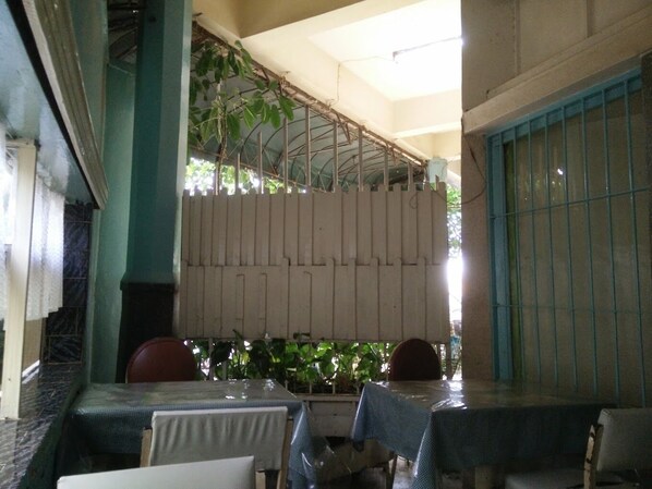 A covered outdoor seating area with tables and chairs, a metal fence, and greenery in the background.