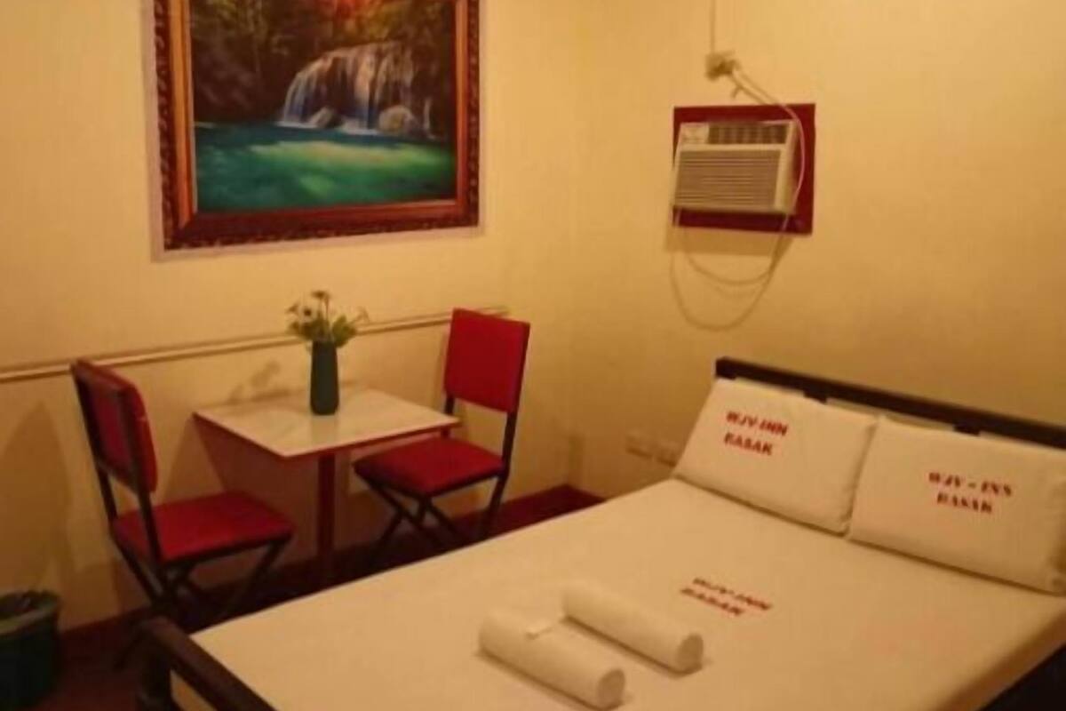A hotel room with a bed, a desk, a chair, a painting of a waterfall, and an air conditioning unit.