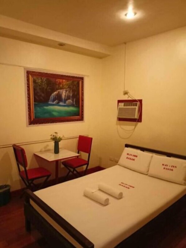 A hotel room with a bed, a desk, a chair, a painting of a waterfall, and an air conditioning unit.
