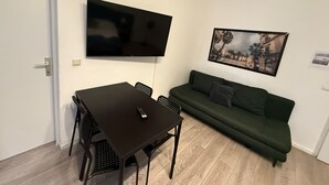Ein kleiner Wohnbereich mit einem dunklen Holztisch, schwarzen Stühlen, einem grünen Sofa, einem fest installierten Flachbildfernseher und einem gerahmten Bild an der Wand.