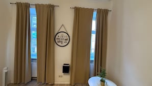 Ein Zimmer mit beige farbenen Vorhängen, einem an der Wand hängenden Bilderrahmen, einem kleinen Tisch mit einer Pflanze und einem Heizkörper.