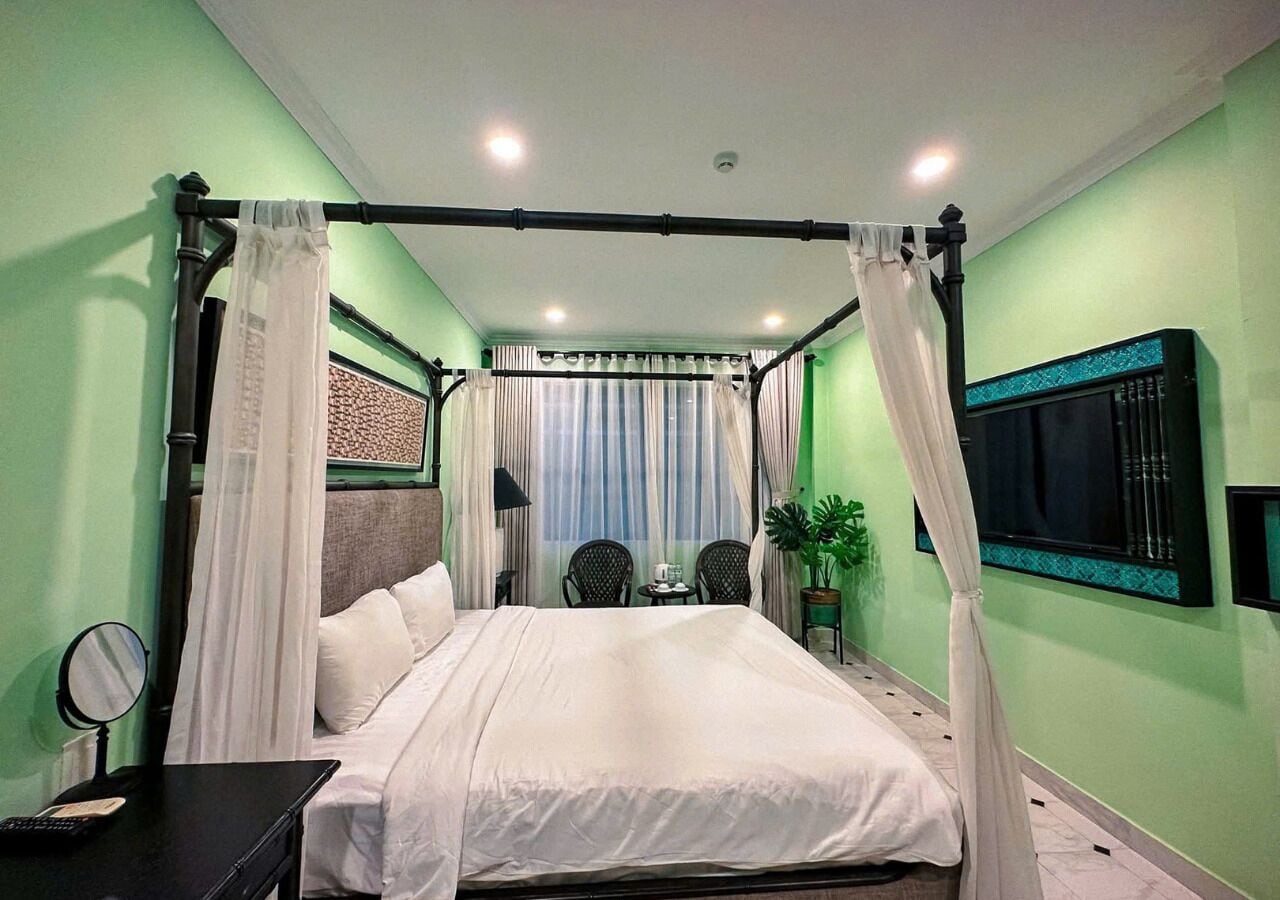 A bedroom with a four-poster bed, a bedside table with a lamp, a small table with chairs, and a potted plant.