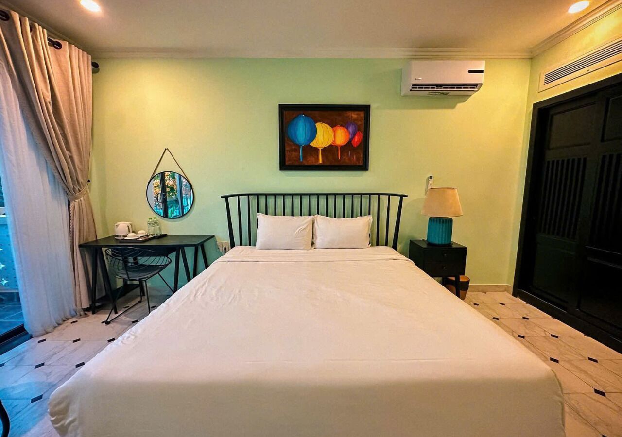 A hotel room with a bed, a desk with a chair, a small table with a lamp, a painting on the wall, and an air conditioning unit.