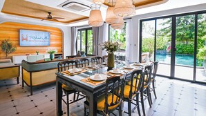 Ein Essbereich mit großem Tisch, gedeckt für eine Mahlzeit, einem Fernseher, der 'Retreat Villa Hometown Signature' zeigt, und Blick auf einen Swimmingpool.