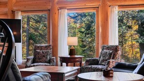 Ein gemütliches Zimmer mit zwei gestreiften Sesseln, einem Holztisch und einer Lampe. Große Fenster bieten Blick auf Herbstbäume.