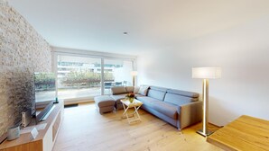 Una sala de estar moderna con un sofá gris, una pequeña mesa de centro y una lámpara de pie. Hay un televisor sobre un mueble de madera y un gran ventanal con vistas al exterior.