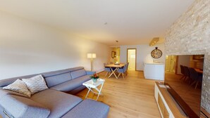 Una sala de estar moderna con un sofá gris, una mesa de comedor con sillas y una cocina visible a través de una puerta abierta.