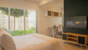 A modern hotel room with a large bed, a desk with a chair, a TV, and a balcony with a view of buildings and greenery. - w* | Duplex Cityview 3BR w/ Balcony in Miraflores (Miraflores)