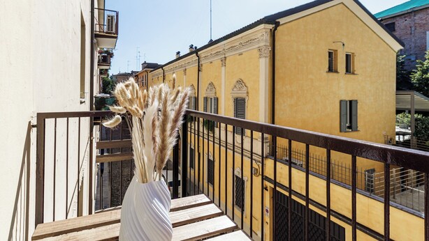 A balcony with a white vase of pampas grass, wooden benches, and a yellow building in the background. - Casa Valentina | WiFi, Train Station, Parking (Modena)