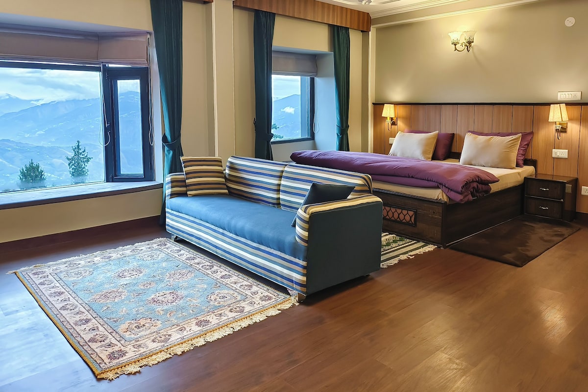 Ein Hotelzimmer mit Sofa, Bett mit Kopfteil, Nachttisch und Blick auf die Berge durch das Fenster.