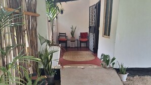 A covered outdoor area with a concrete floor, potted plants, a wooden bench, and a table with a bowl of food.