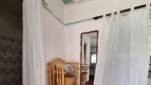 A room with a wooden bed, a small wooden chair, a woven basket, a mirror, and a white curtain.