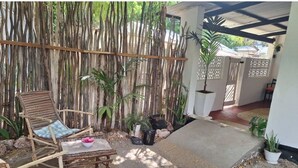 A patio area with wooden furniture, a bamboo fence, potted plants, and a concrete pathway.