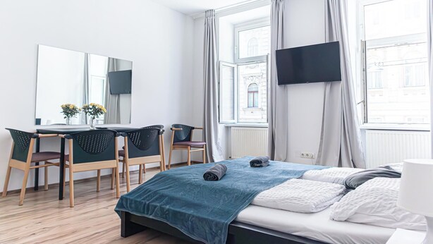Moderni hotellihuone, jossa on sänky, työpöytä tuoleineen, taulutelevisio ja suuret ikkunat, joissa on verhot.
