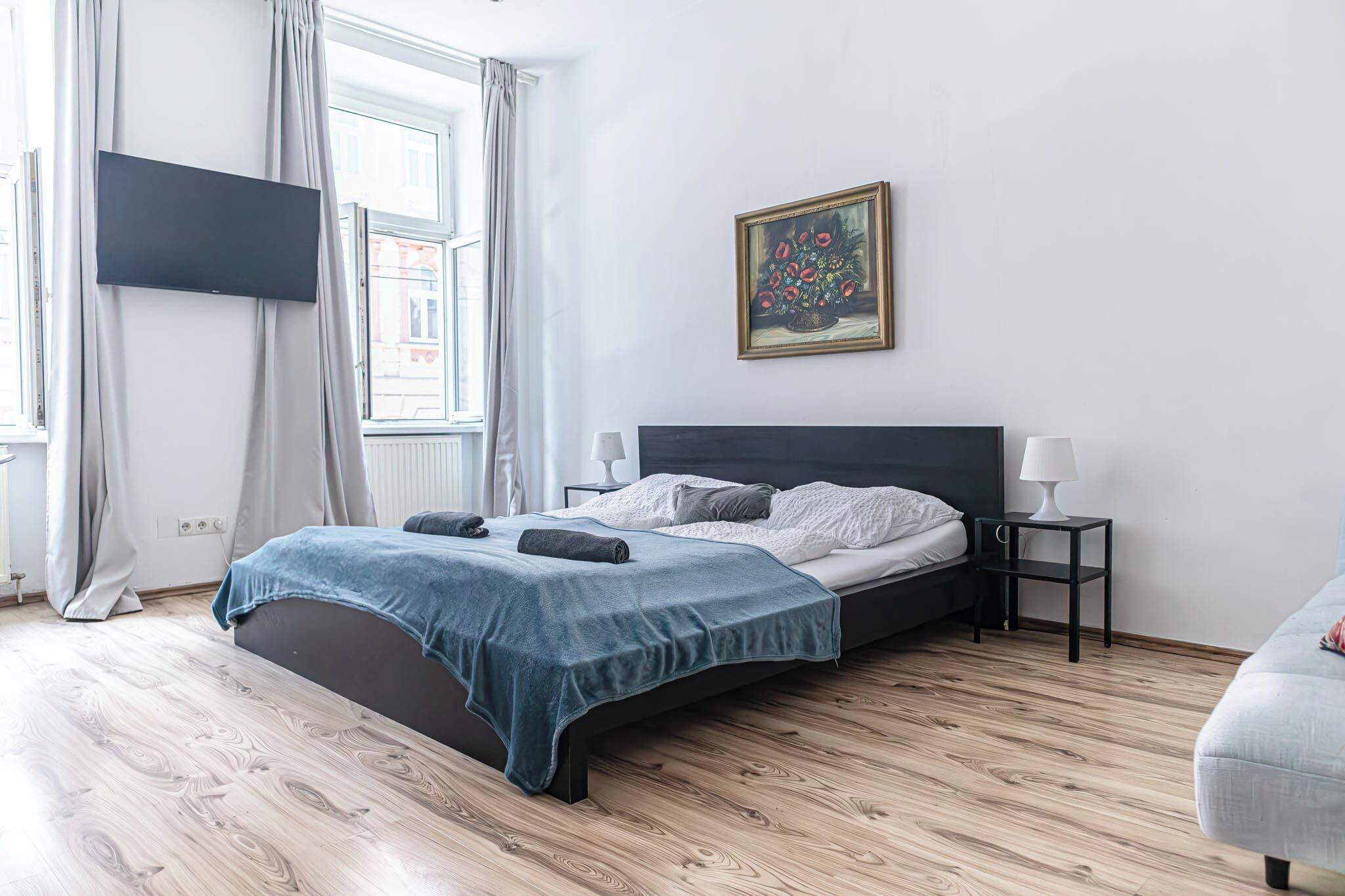 Un dormitorio con cama, televisor montado en la pared, un cuadro y una ventana con cortinas.