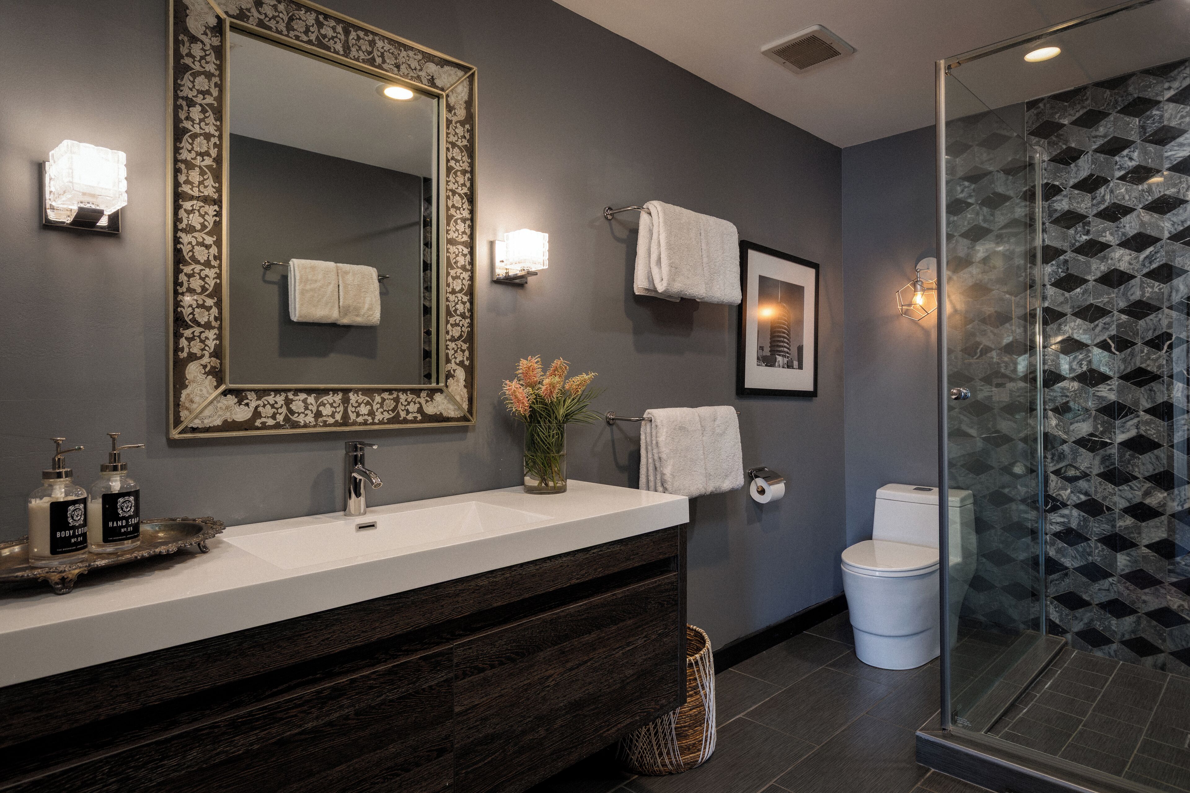 A modern bathroom with a large mirror, a white sink, dark wood vanity, two hand soap dispensers, a vase with flowers, a toilet, and a shower with a geometric tile backsplash.