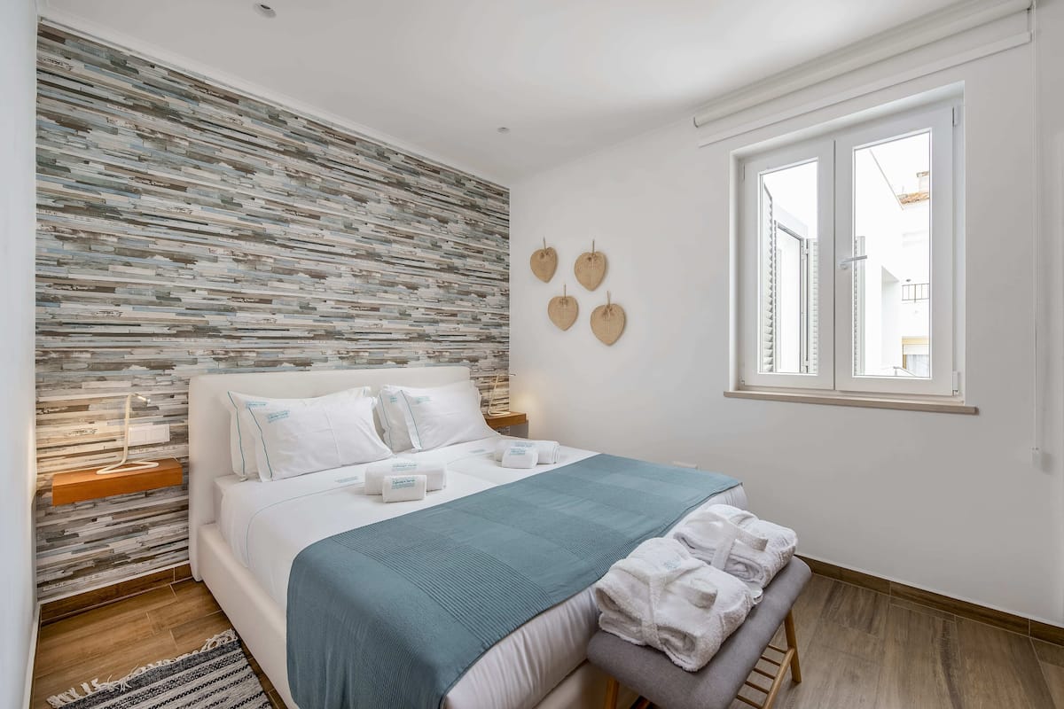 Un dormitorio ordenado con cama, mesitas de noche, ventana y pared con textura.