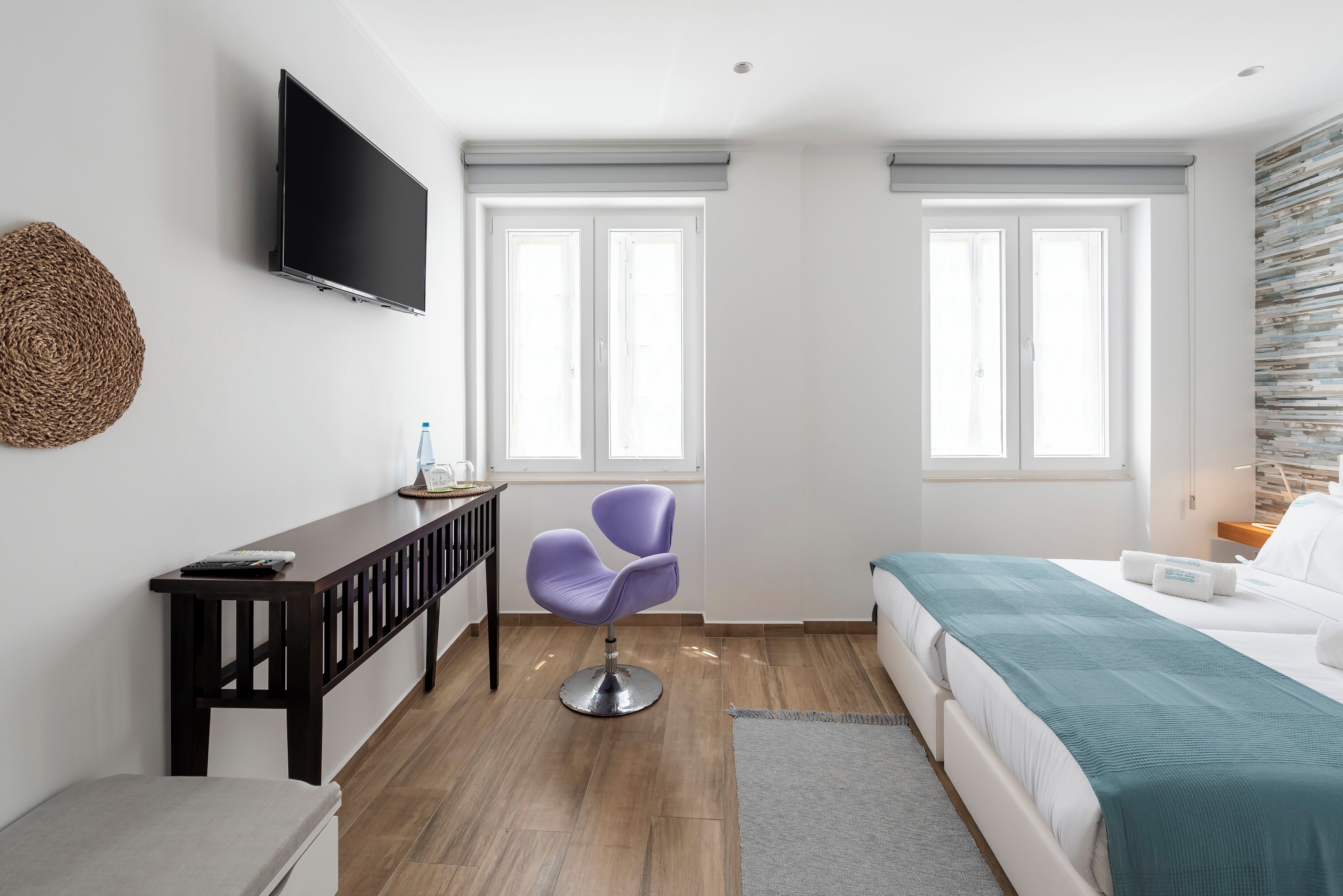 A modern hotel room with a bed, a desk with a chair, a TV, and two windows.