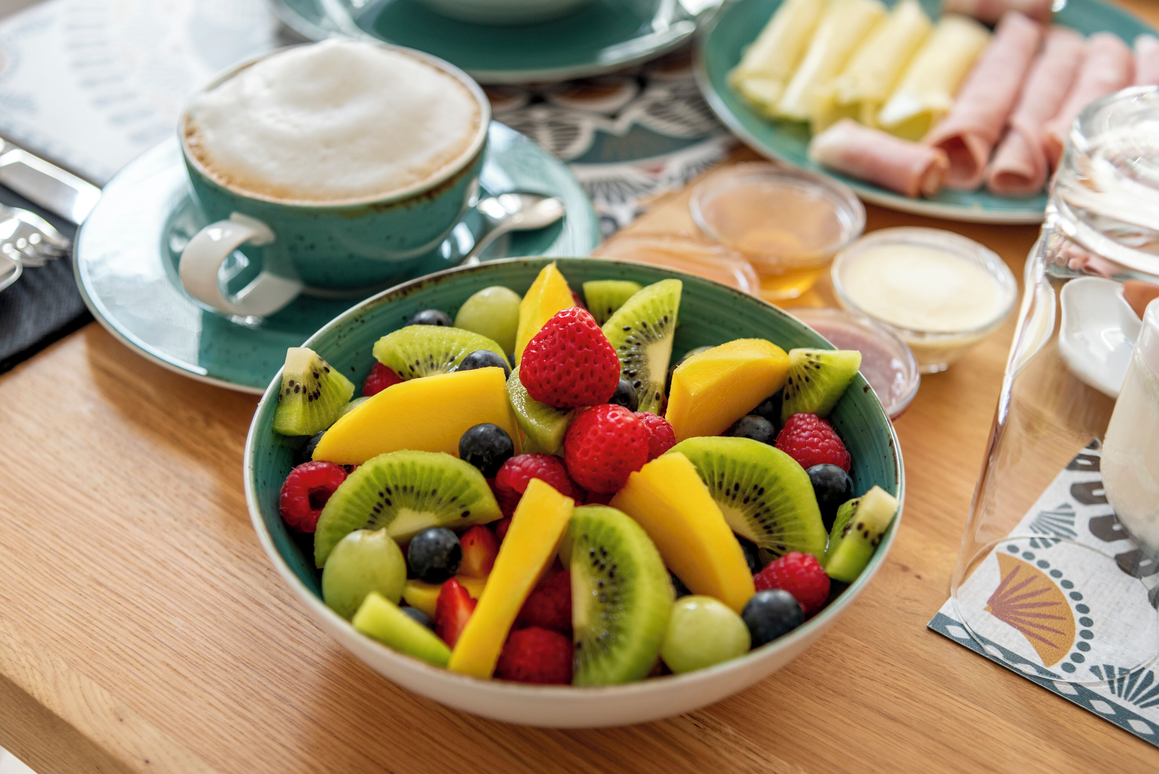Un tazón de ensalada de frutas mixtas sobre una mesa, con una taza de café y platos de comida al fondo.