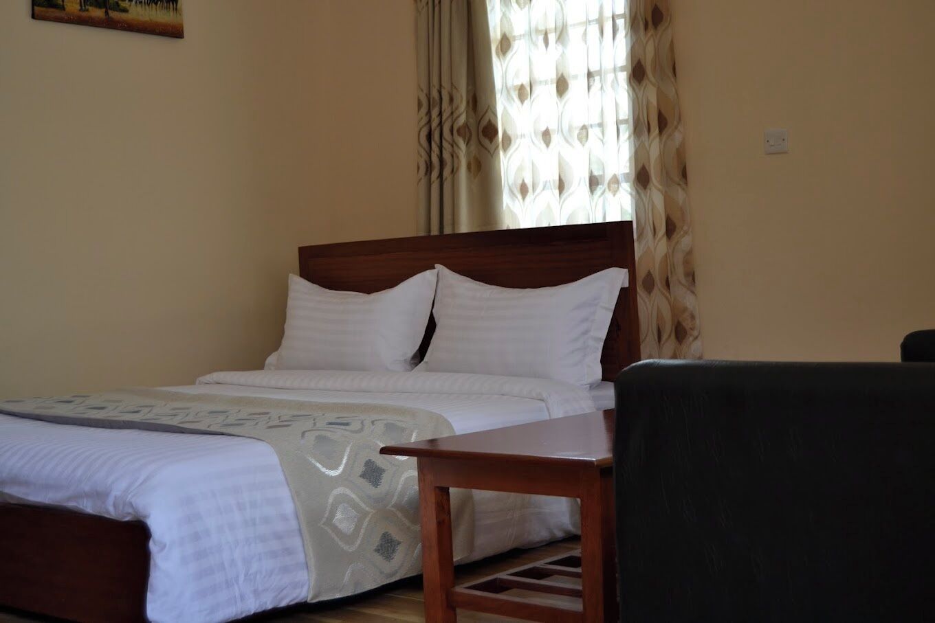 A neatly made bed with white linens and a patterned throw, a wooden nightstand, and a black chair in a room with beige walls and a window with patterned curtains.
